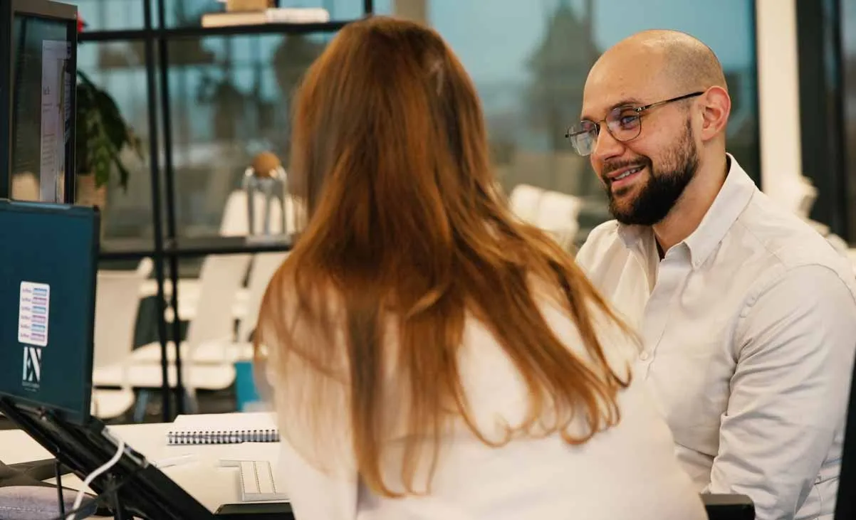 A team member smiling while talking with a colleague at their desk